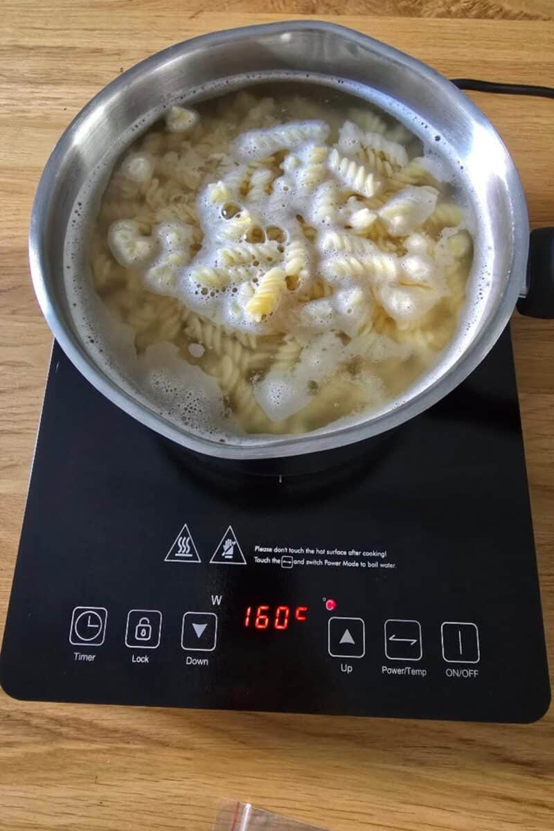 A pot of fusilli pasta boils on the AMZCHEF Single Induction Hob, set at 160°C for compact convenience. Foam forms on top as the cooktop displays various buttons and the temperature in bold red numbers.