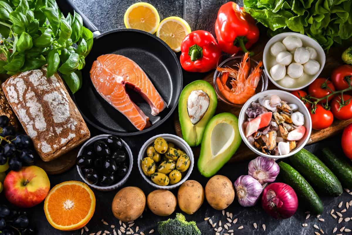 A variety of fresh foods inspired by the Nordic Diet, including salmon, shrimp, avocado, bread, olives, potatoes, cheese, peppers, greens, fruits, garlic, onion, nuts, seeds, and lemon arranged on a table.