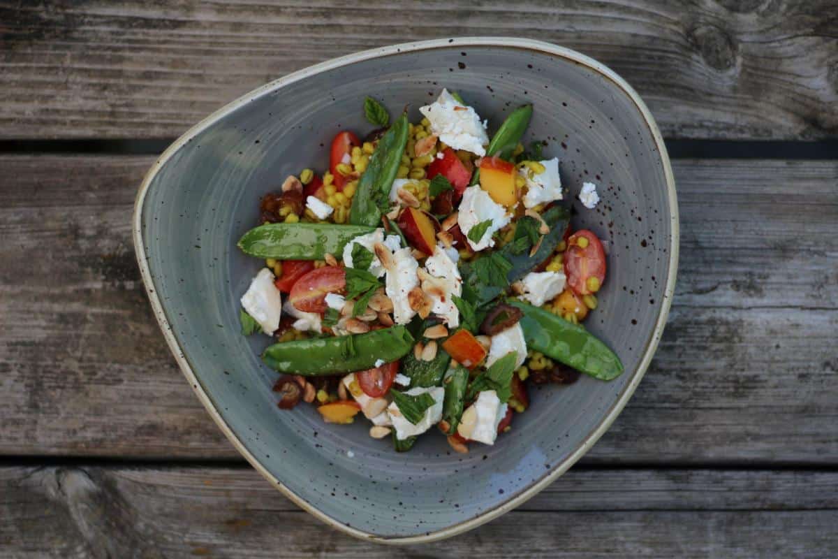 A vibrant summer barley grain bowl with snap peas, cherry tomatoes, sweetcorn, crumbled cheese, chopped nuts, and fresh herbs on a rustic wooden table—perfect for better health.