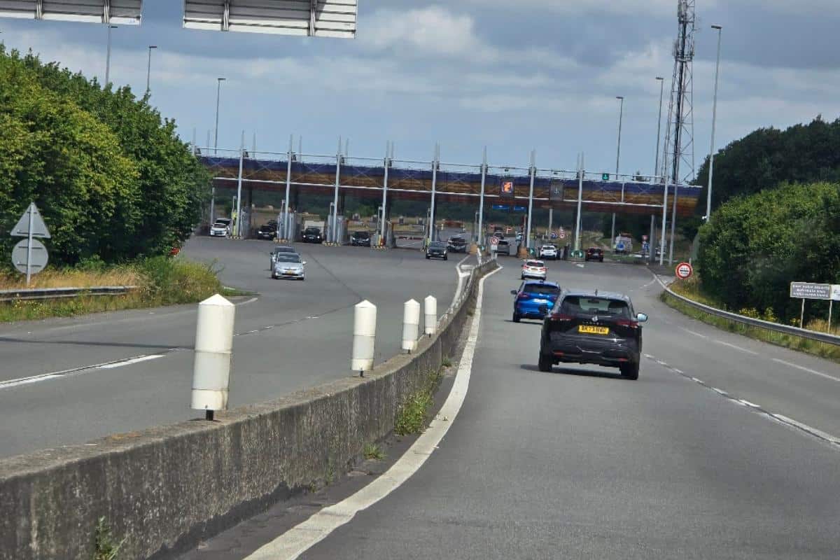 Cars drive on a divided French toll road with a concrete barrier, surrounded by green trees under a blue sky. An overpass and traffic signs are visible in the background—perfect for any Emovis tag guide.