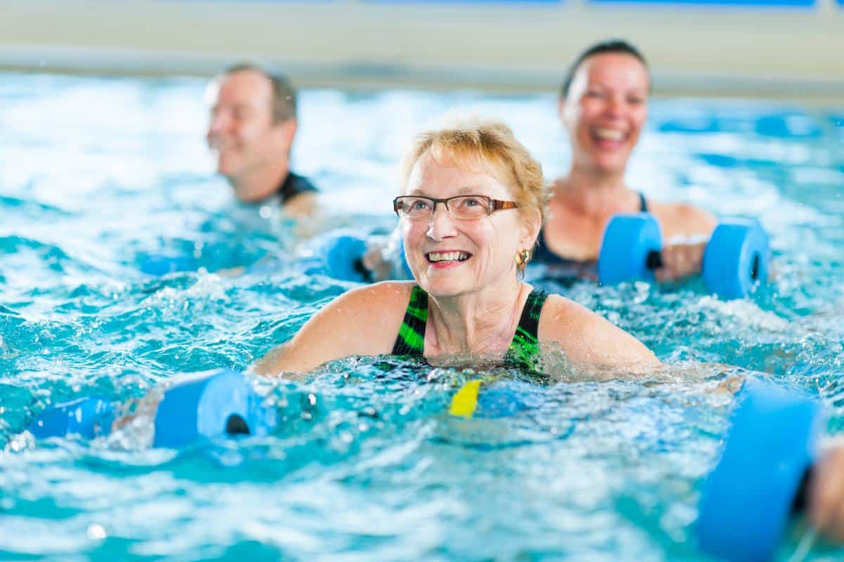 Three adults, including a smiling older woman in glasses, participate in a water exercise class focused on exercises for bad knees, each holding blue foam dumbbells. Other participants are visible in the background.