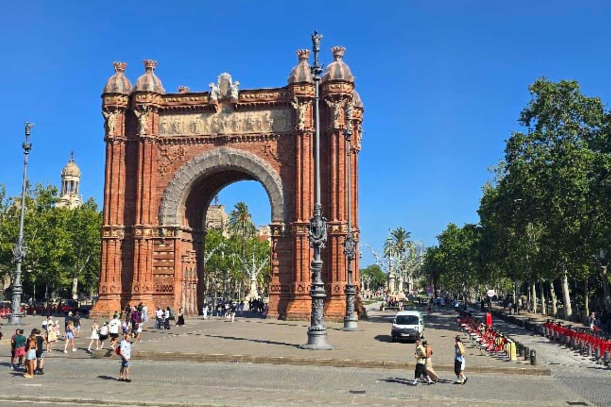 The Arc de Triomf, a large red-brick triumphal arch in Barcelona, stands under a clear blue sky, surrounded by trees and people walking and cycling—an ideal spot to pause during your Barcelona city tour bus adventure.