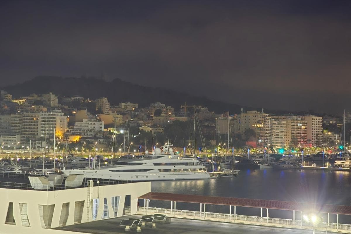 A marina at night with a large white yacht docked in the water, surrounded by smaller boats—reminiscent of an overnight journey from Barcelona to Palma. City buildings and hills glow in the background under a cloudy sky.