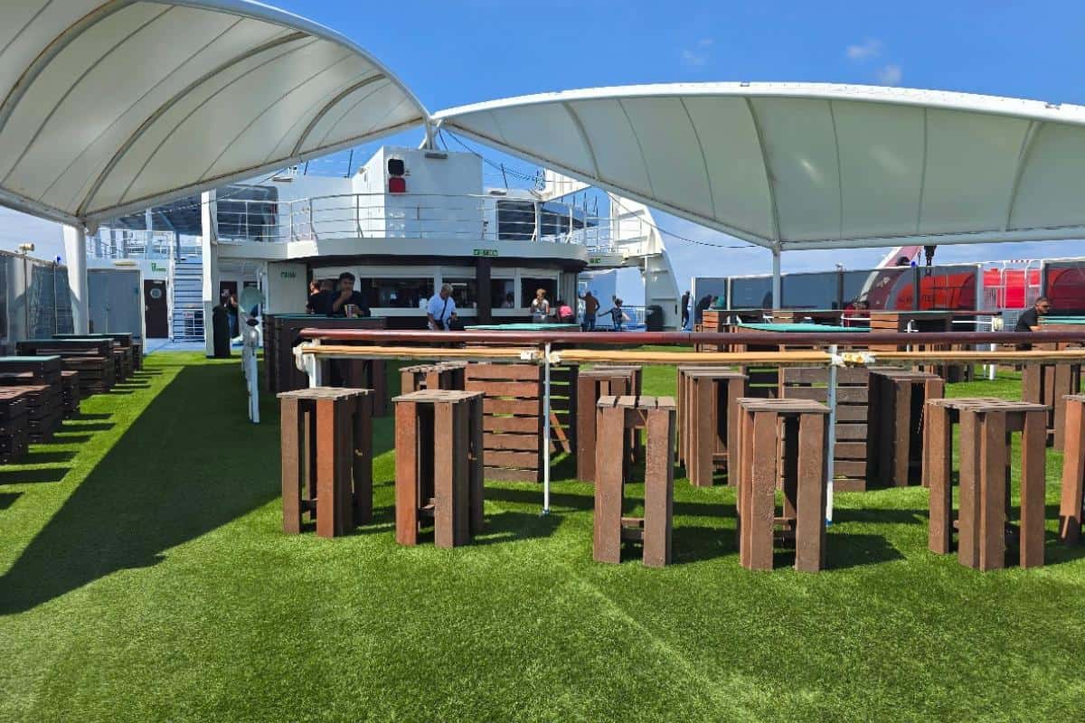 Outdoor seating area on a Balearia Ferry with artificial grass, wooden tables and benches, and white canopies overhead. People are gathered near the ship’s railings under a sunny blue sky on the Alcudia to Barcelona route.