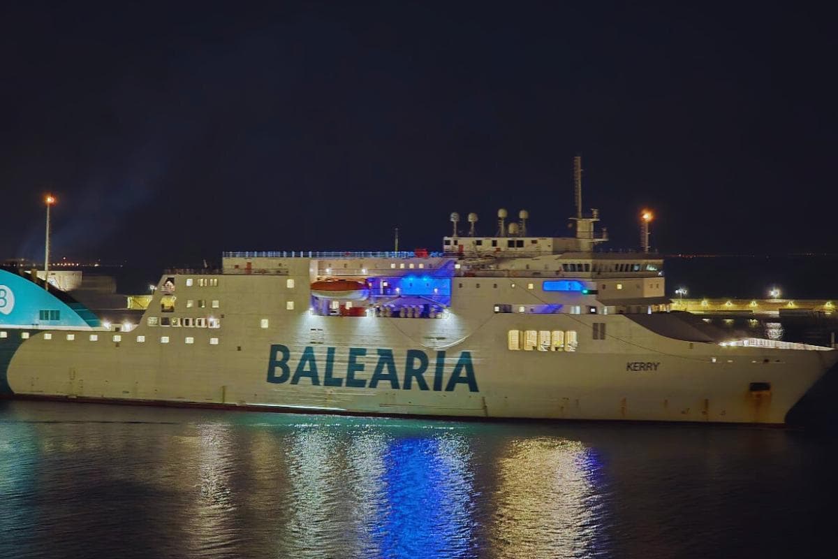 A large white Balearia Ferry with "BALEARIA" written on the side is illuminated at night, reflecting lights onto calm water. The ship's name, "KERRY," near the bow, suggests a possible Alcudia to Barcelona route.