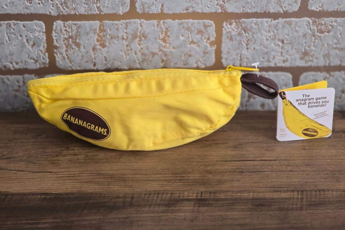A yellow, banana-shaped zippered pouch labeled "Bananagrams" rests on a wooden surface in front of a brick wall. An attached tag describes it as an anagram game—one of the best family games for holiday fun and lightweight family game nights.