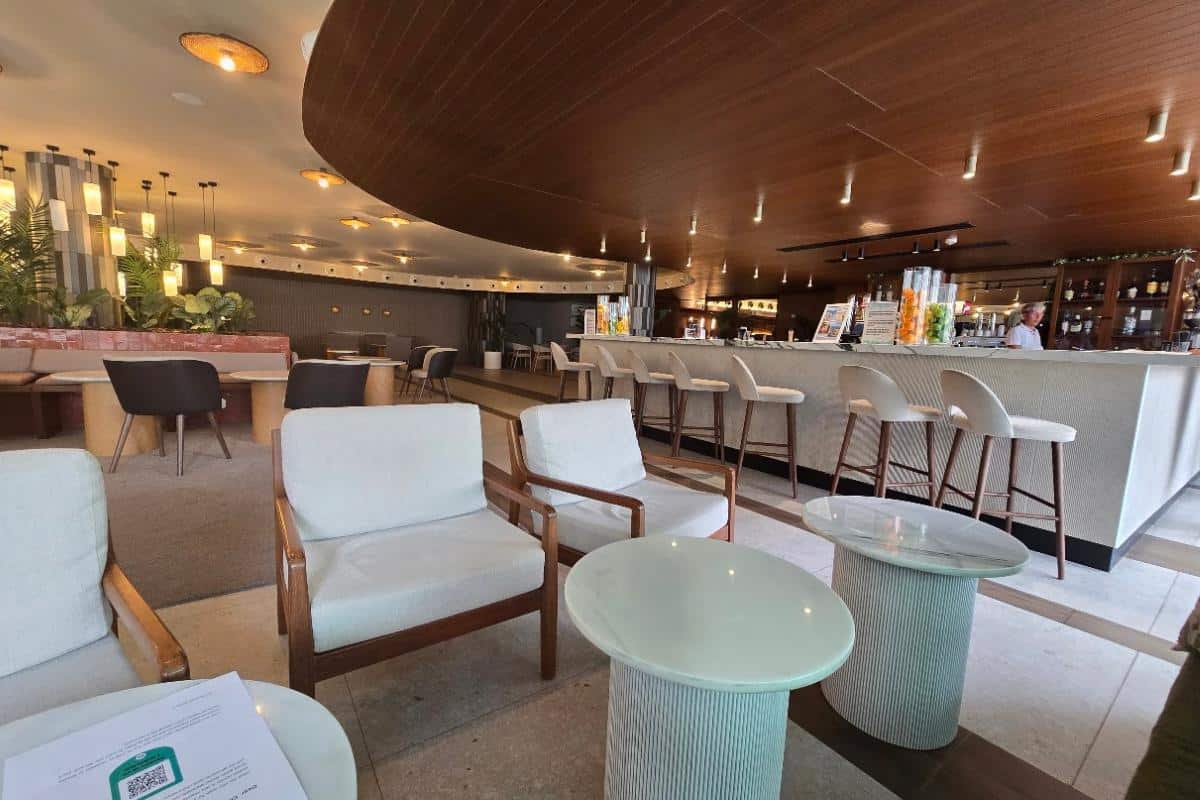 Modern hotel lounge at Bahia del Sol in Santa Ponsa, featuring white cushioned chairs, round tables, and a long bar counter with high stools. Warm lighting and wood-paneled ceiling create a cozy, upscale vibe—perfect for an all-inclusive getaway.