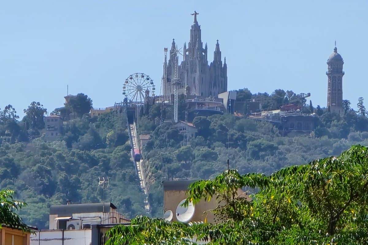 A distant view of Tibidabo hill in Barcelona, featuring the Temple Expiatori del Sagrat Cor church, a ferris wheel, a tower, green trees, and several buildings—perfect for families exploring by City Tour Bus under a clear sky.