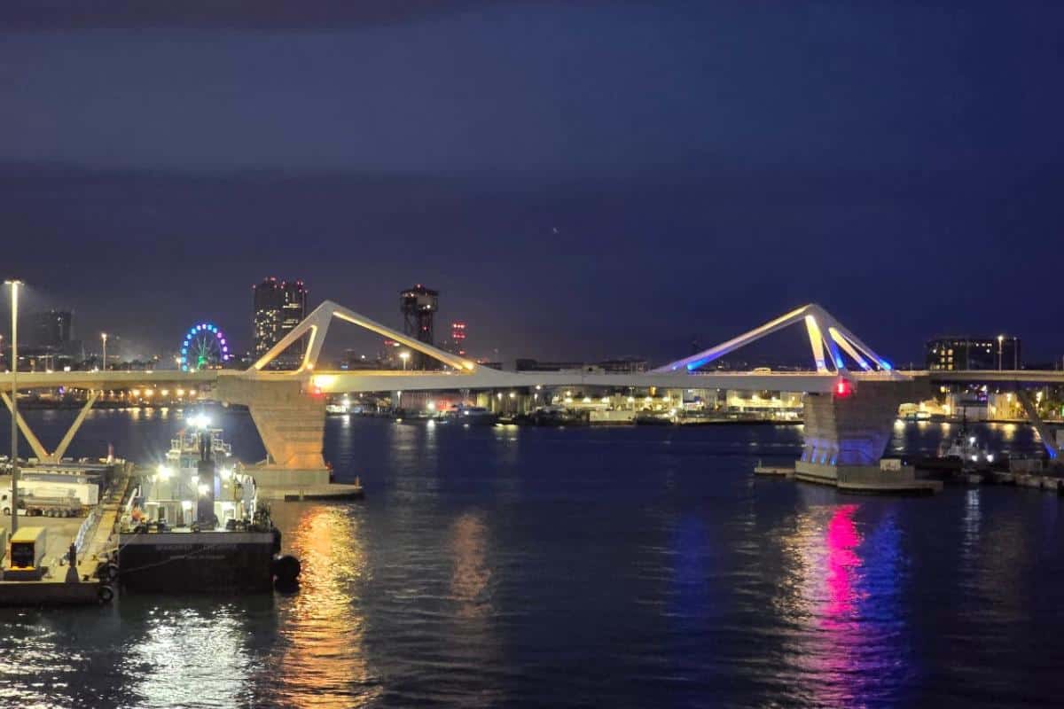 A modern, illuminated bridge spans across a river at night, with colorful lights reflecting on the water—much like the dazzling views you might enjoy on an overnight journey from Barcelona to Palma with GNV Ferries. City buildings and a lit Ferris wheel glow in the background.