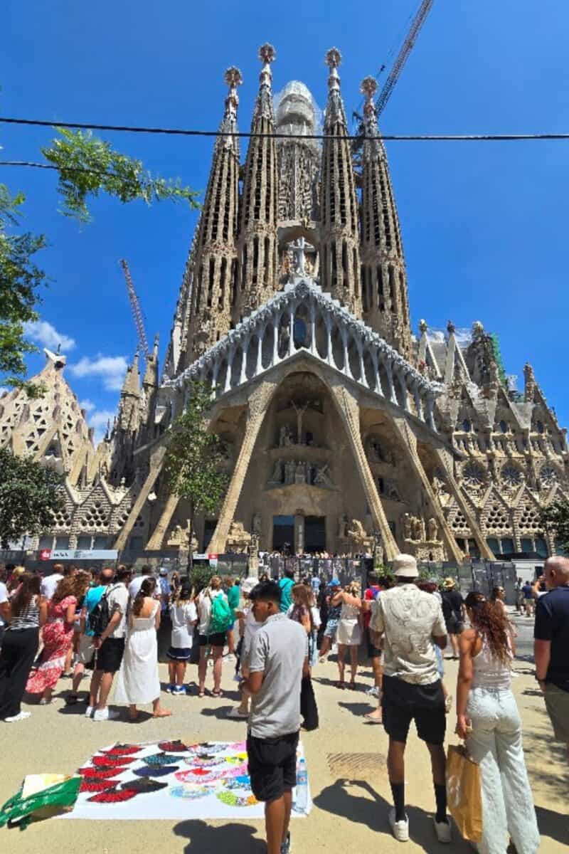 Families gather in front of the Sagrada Família basilica in Barcelona under a blue sky, with street vendors selling colorful items nearby. Tall, detailed towers and cranes rise above the cathedral, while a City Tour Bus passes in the background.