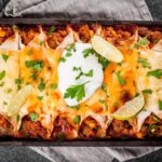 A casserole dish filled with Easy Enchiladas topped with melted cheese, sliced lime wedges, a dollop of sour cream, and fresh cilantro. The dish sits on a gray napkin on a dark countertop.
