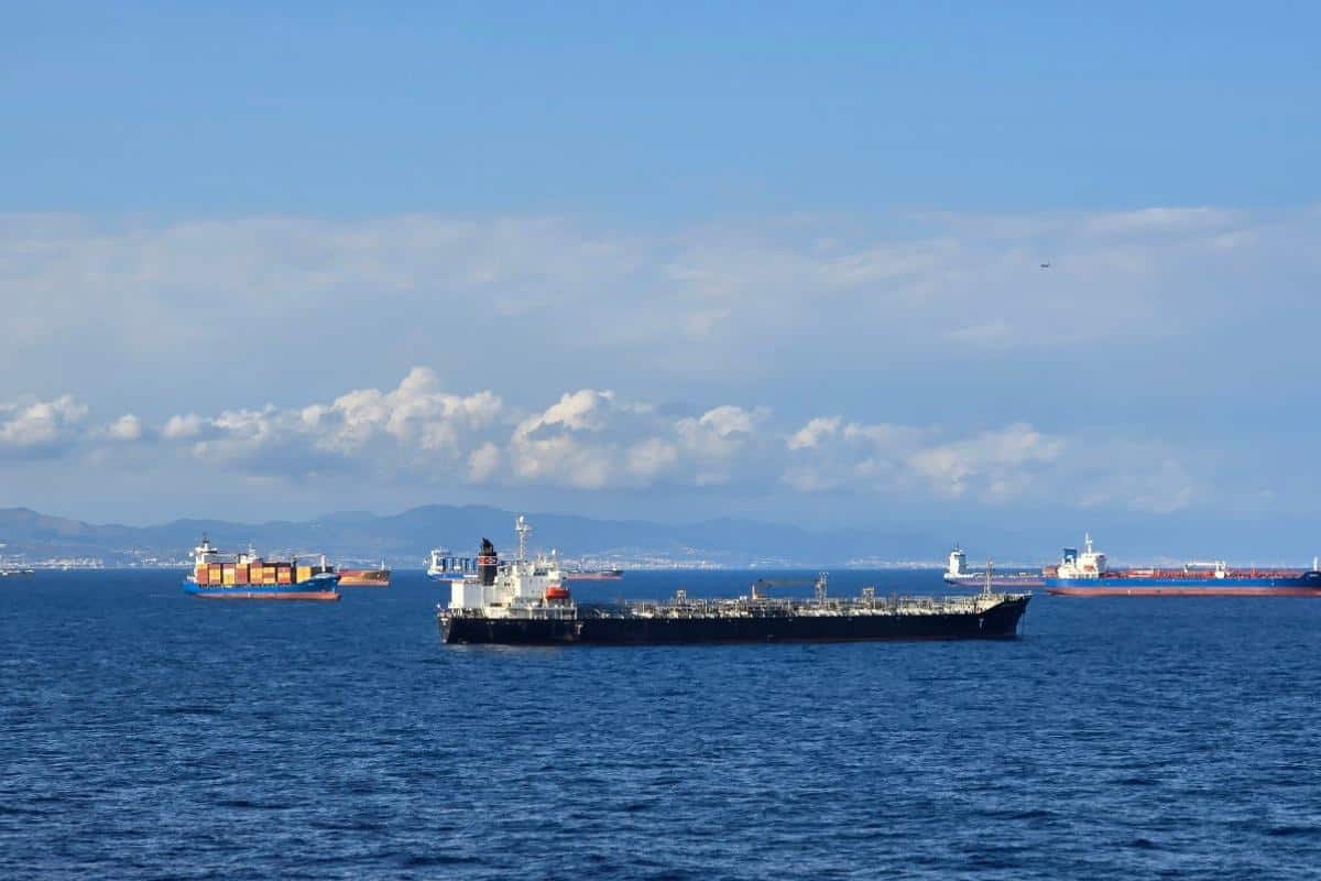 Several large cargo ships, tankers, and a Balearia Ferry are anchored on calm blue water under a partly cloudy sky, with distant mountains visible on the horizon—reminiscent of scenic views on the Alcudia to Barcelona route.