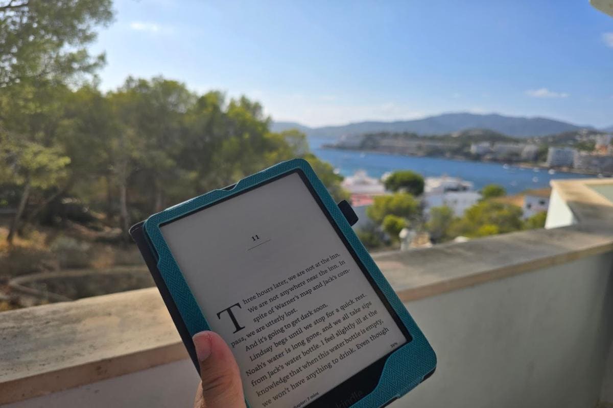 A person enjoys a Midweek Catch-Up, holding an e-reader displaying a book against a scenic outdoor view of trees, water, boats, and distant hills under a clear blue sky.