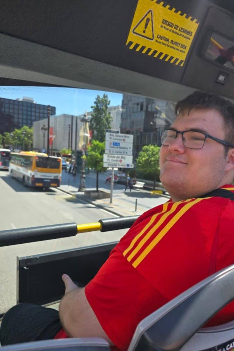 A man in a red shirt with yellow stripes sits on the upper deck of a Barcelona city tour bus, smiling at the camera. City buildings, buses, and a street sign are visible in the background on a sunny day.