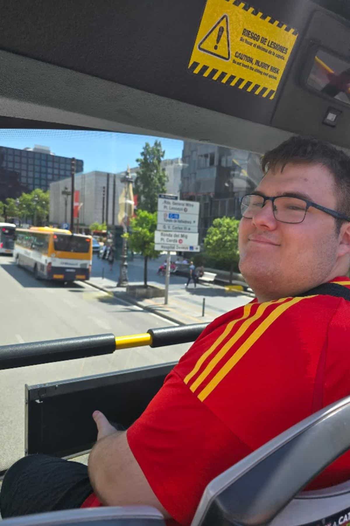 A man in a red shirt with yellow stripes sits on the upper deck of a Barcelona city tour bus, smiling at the camera. City buildings, buses, and a street sign are visible in the background on a sunny day.