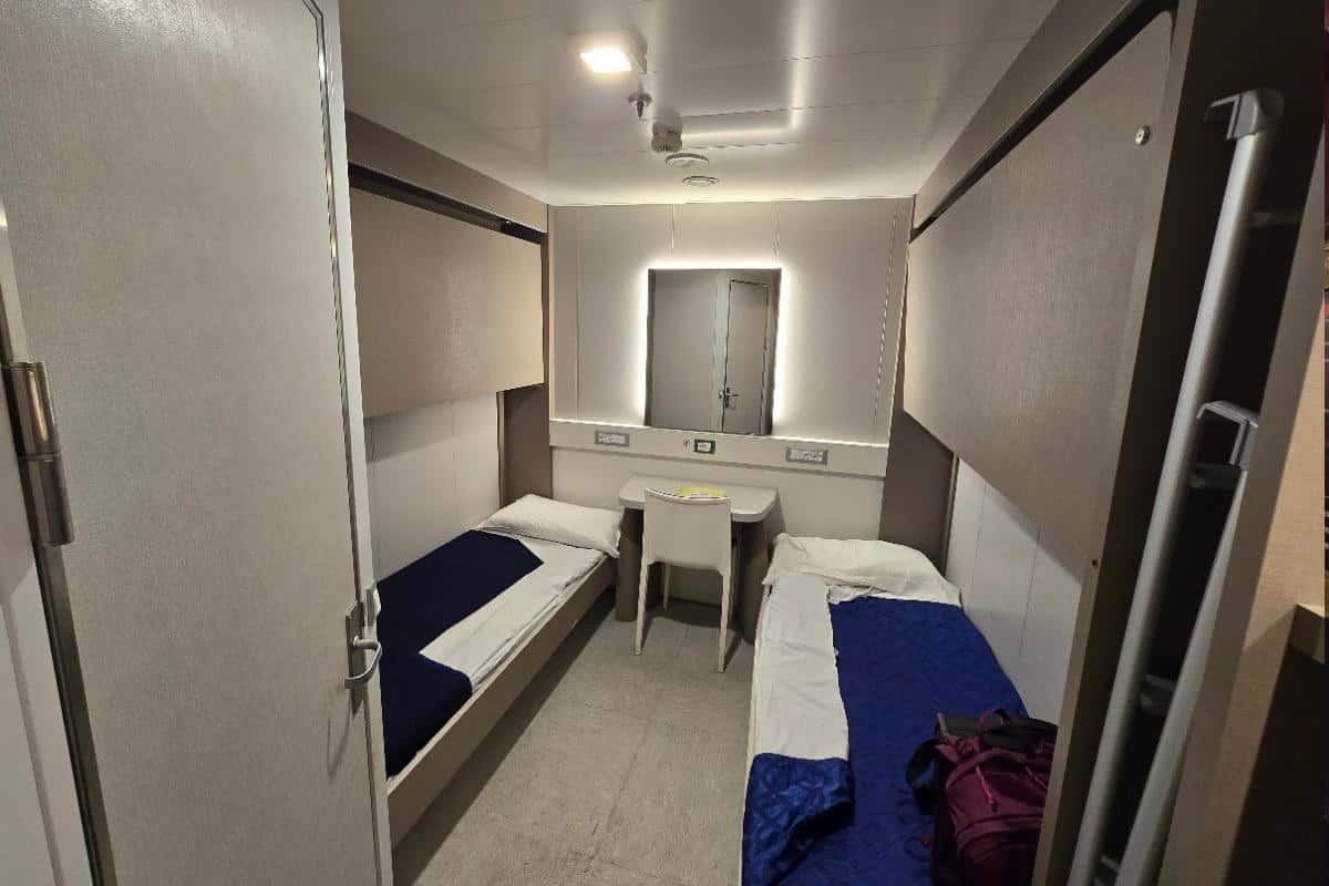A small, modern cabin on GNV Ferries’ Barcelona to Palma route features two single beds, a small table with a mirror, overhead shelves, and a suitcase on one bed. The neutral tones and bright ceiling lights ensure comfort for your overnight journey.
