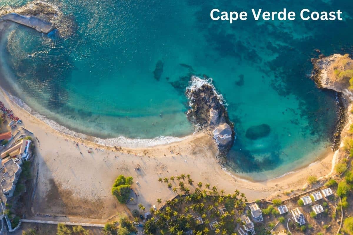 Aerial view of a sandy beach on the Cape Verde coast with turquoise water, lush palm trees, and shoreline buildings. Perfect for first-time visitors, this travel guide highlights the scenic curve around a rocky area. Text reads "Cape Verde Coast.