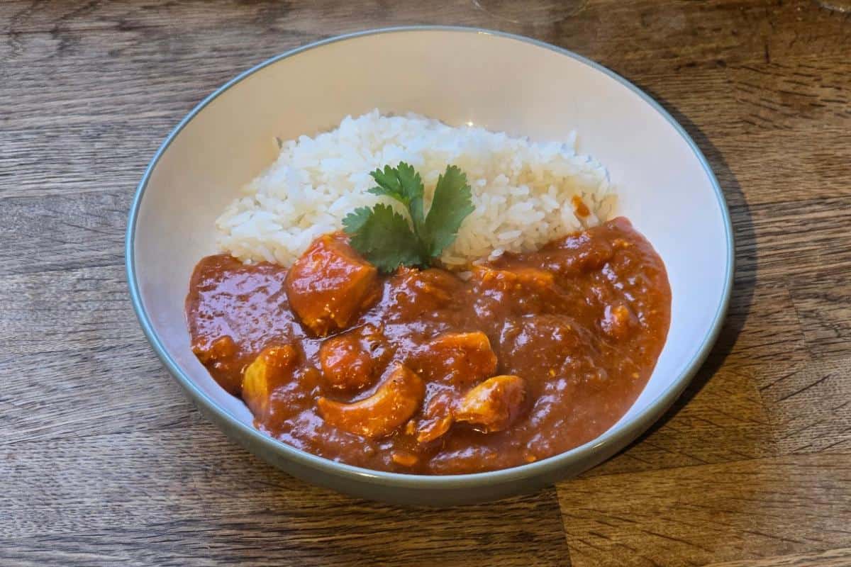 A bowl of white rice and red curry with chicken pieces, inspired by a classic chicken bhuna recipe, garnished with cilantro and served on a wooden surface.
