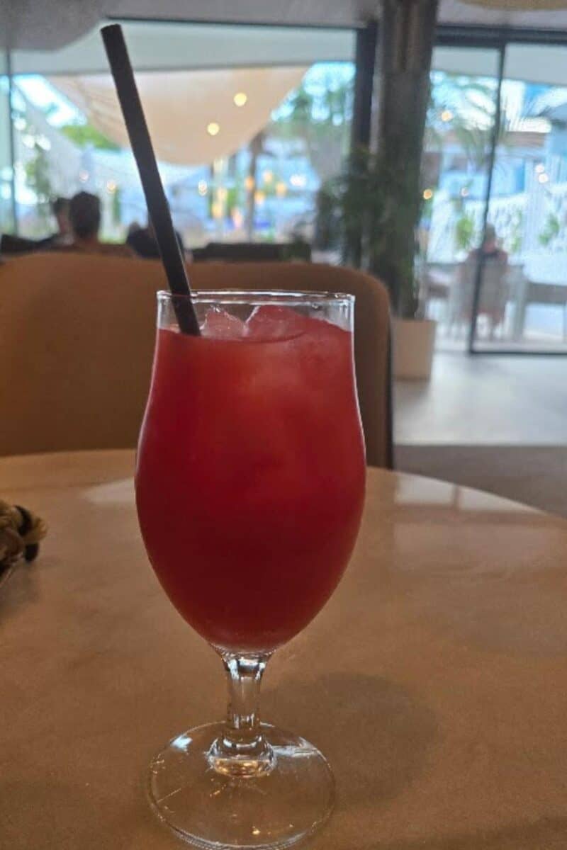 A glass of red iced drink with a black straw sits on a table in a modern indoor café at Bahia del Sol, with blurred background showing plants, large windows, and soft lighting—perfect for an all-inclusive Santa Ponsa escape.