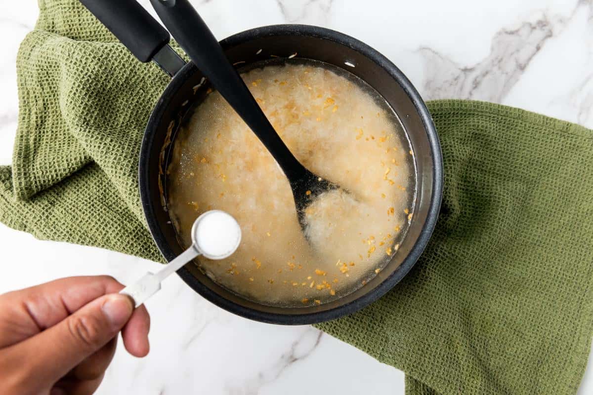 A hand adds a spoonful of salt to a saucepan of easy garlic rice, with rice and water simmering together. The pot, on a green kitchen towel atop marble, is being stirred with a black spoon—perfect as a flavorful side dish.
