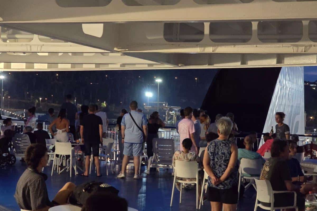 A group of people, some seated and some standing, gather on the deck of a GNV Ferries ship during an overnight journey from Barcelona to Palma. White chairs and tables line the deck as city lights twinkle in the background.