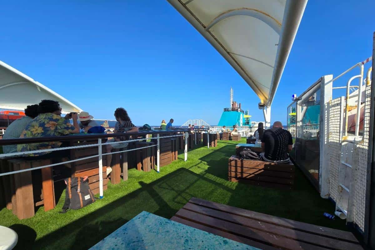 Passengers relax on benches and tables on the deck of a Balearia Ferry under a bright blue sky, with artificial grass flooring and ship structures in the background—an inviting scene on the Alcudia to Barcelona route.