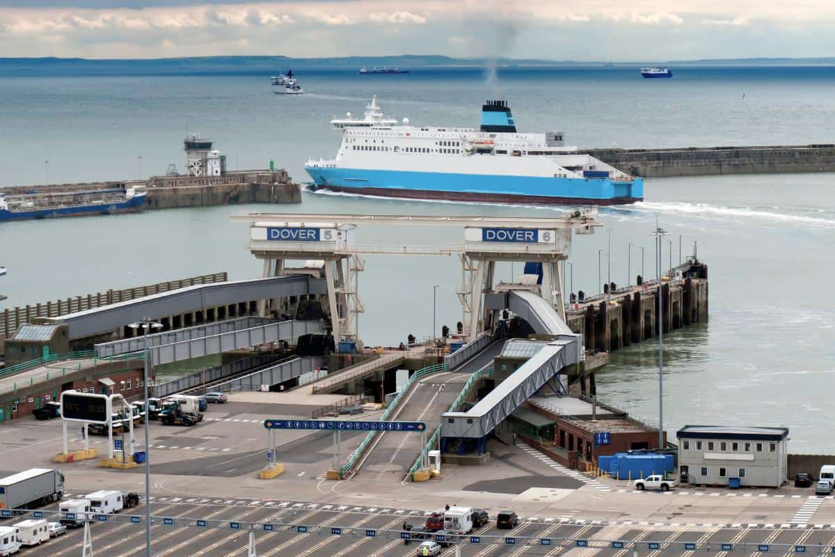 A large Dover to Calais Ferry approaches the dock at Dover Port, with ramps, terminals, and vehicles visible in the foreground, and the sea and other ships in the background under a cloudy sky.
