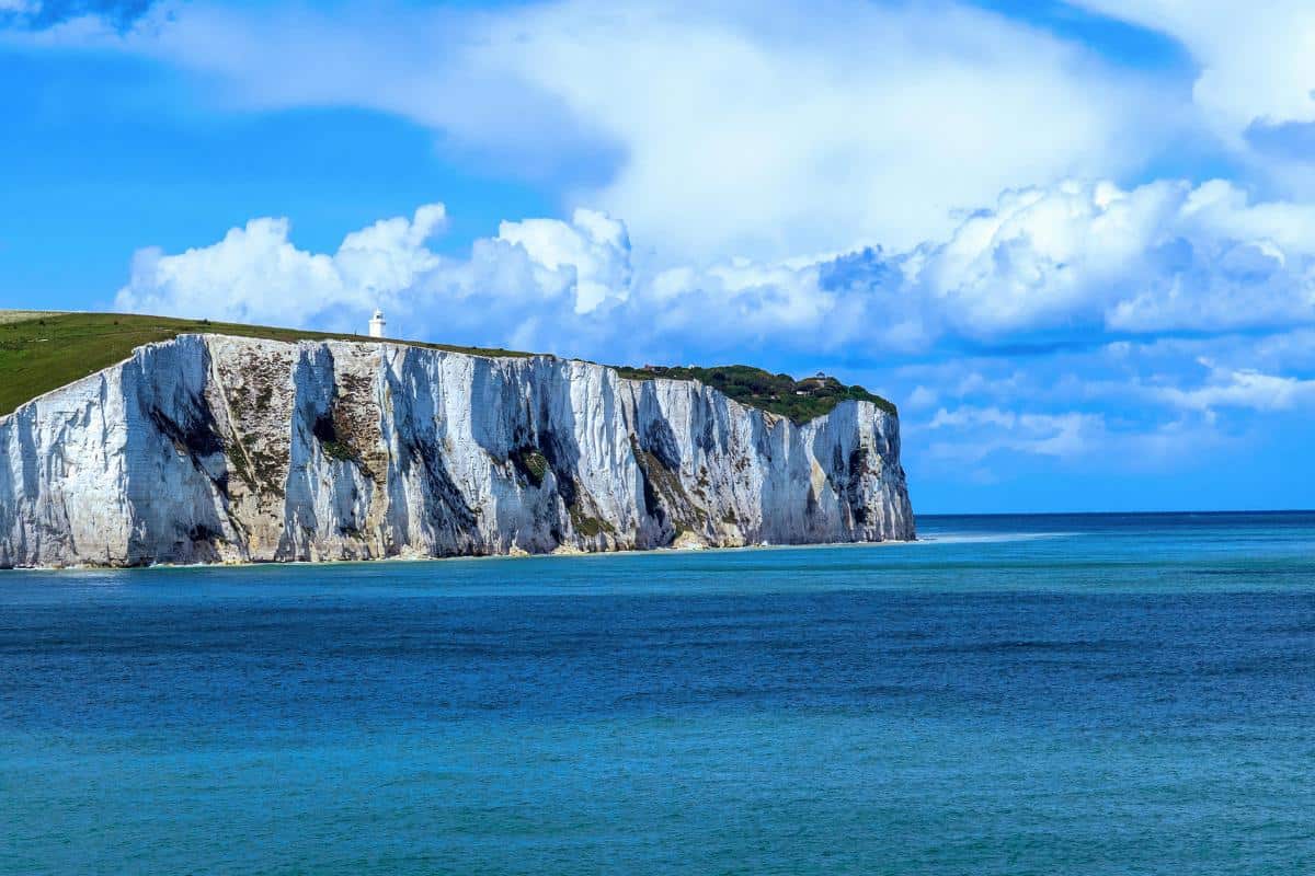 White chalk cliffs rise above the blue sea, topped with green grass and a distant white lighthouse under a partly cloudy sky—a classic view often seen on the Dover to Calais Ferry, perfect for any travel guide.