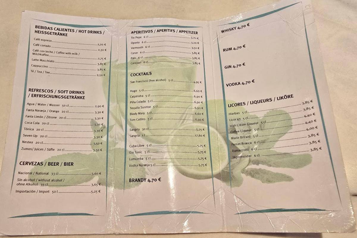 A trifold restaurant menu at Bahia del Sol in Santa Ponsa lists hot drinks, soft drinks, beer, and more in Spanish, German, and English with prices in euros. The green leaf background completes the all-inclusive dining experience.