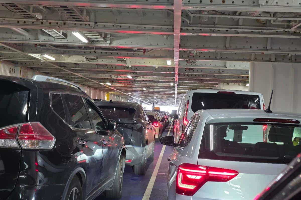 Rows of cars are parked closely together inside the lower deck of the Dover to Calais Ferry, with overhead metal beams and fluorescent lights on the ceiling. Red brake lights illuminate the scene, indicating vehicles are stationary.