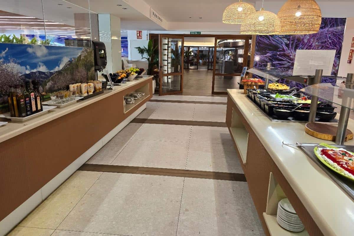 A buffet setup in the modern dining area at Bahia del Sol, Santa Ponsa, features various salads, oils, sauces, fruits, and cold dishes arranged neatly on long counters with glass covers and decorative hanging lights overhead.