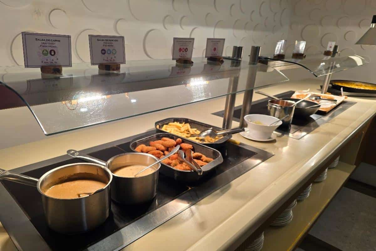 An all-inclusive buffet counter at Bahia del Sol in Santa Ponsa displays trays of food—fries, breaded items, sauces in saucepans, and a bowl—under a glass sneeze guard with food labels. Plates are stacked on shelves below the counter.