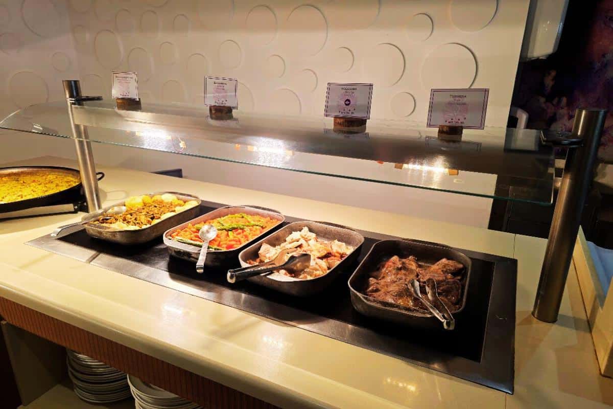 An all-inclusive buffet counter at Bahia del Sol, Santa Ponsa, features five trays of hot food—including meat, vegetables, and pasta—under a glass sneeze guard, with menu cards displayed behind and plates stacked below.