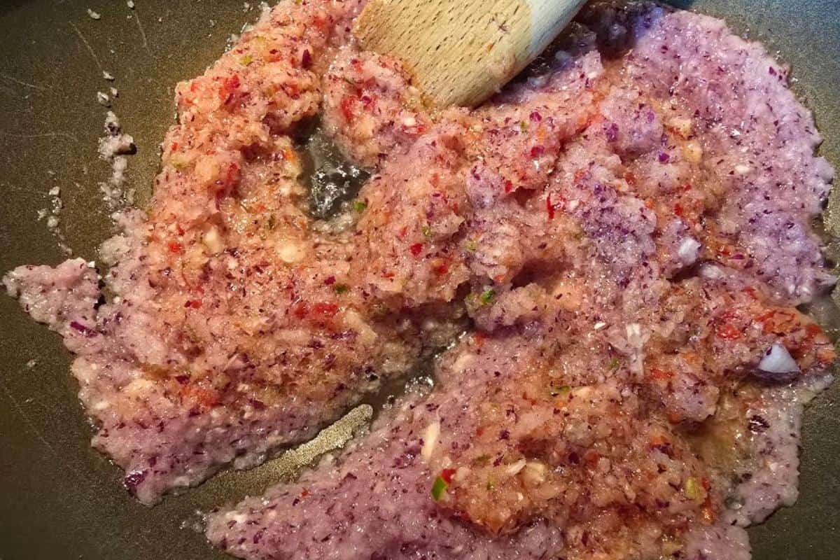 Finely blended mixture of red onions, garlic, chilies, and other ingredients for a chicken bhuna recipe is being sautéed in a pan. A wooden spatula stirs the colorful, textured paste as it cooks.