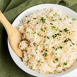 A white bowl filled with cooked Garlic Rice garnished with chopped herbs sits on a green textured cloth, with a wooden spoon resting inside the bowl—an easy recipe perfect as a flavorful side dish.