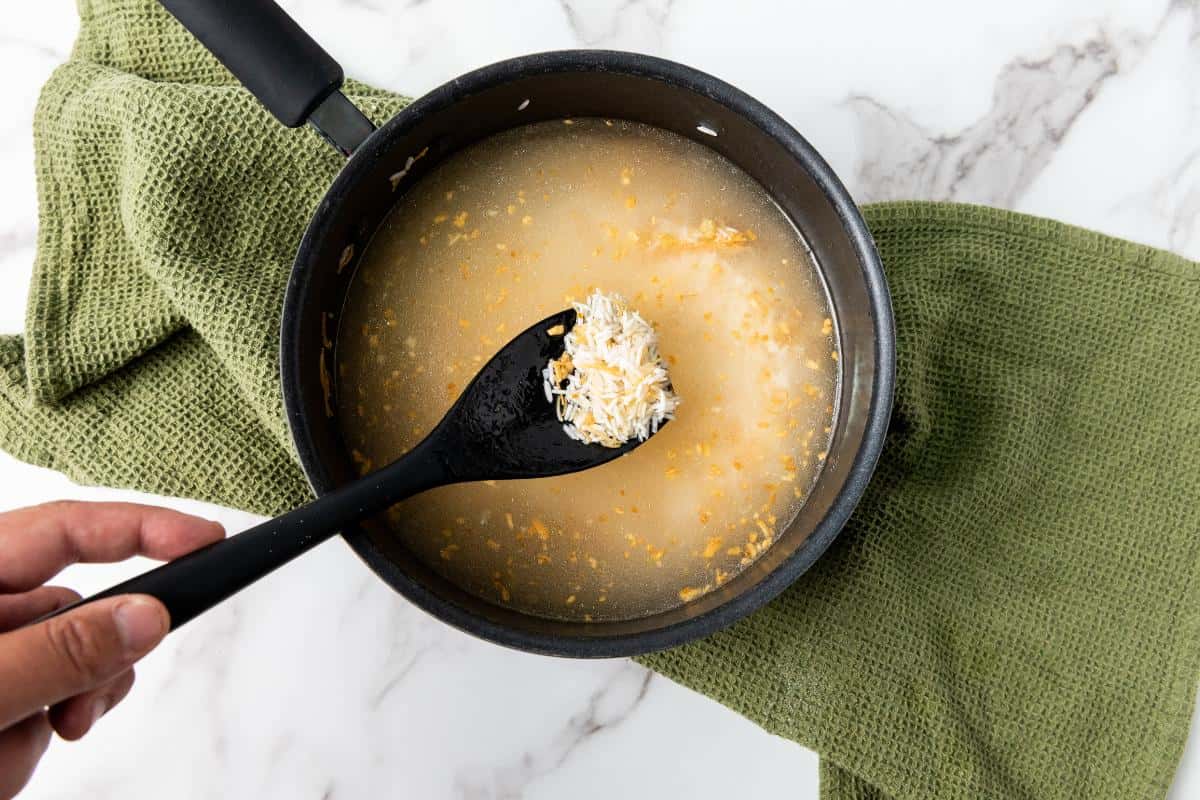 A hand holds a black spoon over a saucepan filled with water and shredded cheese, perfect for preparing delicious rice or an easy side dish, resting on a green kitchen towel on a white marble surface.