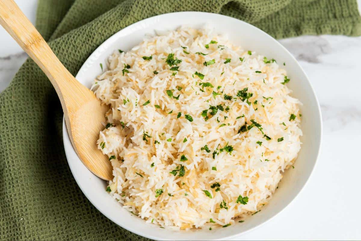 A white bowl filled with cooked Garlic Rice garnished with chopped herbs sits on a green textured cloth, with a wooden spoon resting inside the bowl—an easy recipe perfect as a flavorful side dish.