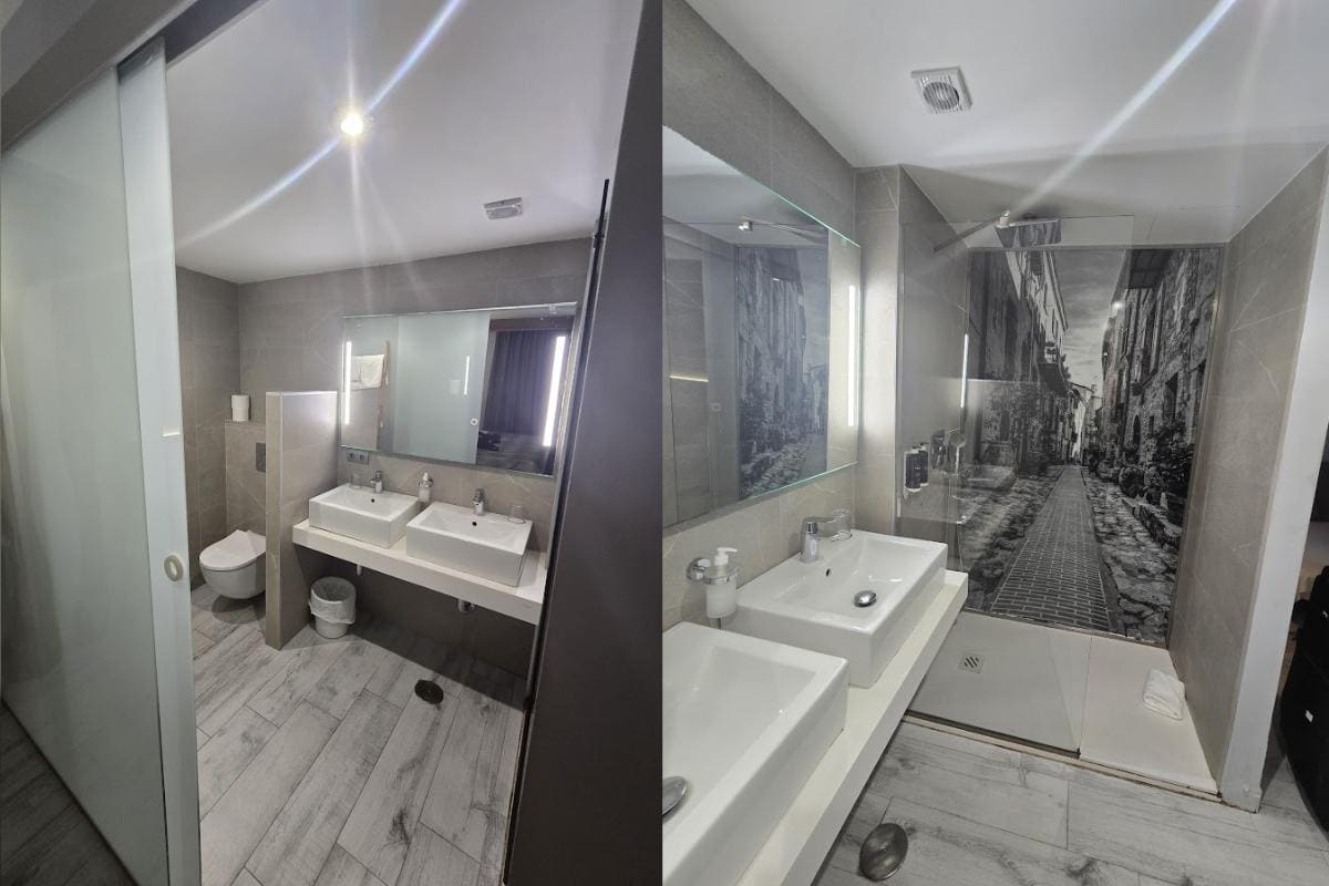 Two photos of a modern bathroom with gray tiles at Bahia del Sol, featuring a city street mural in the glass shower, a double sink vanity with large mirrors, and a toilet partially separated by a frosted glass partition.