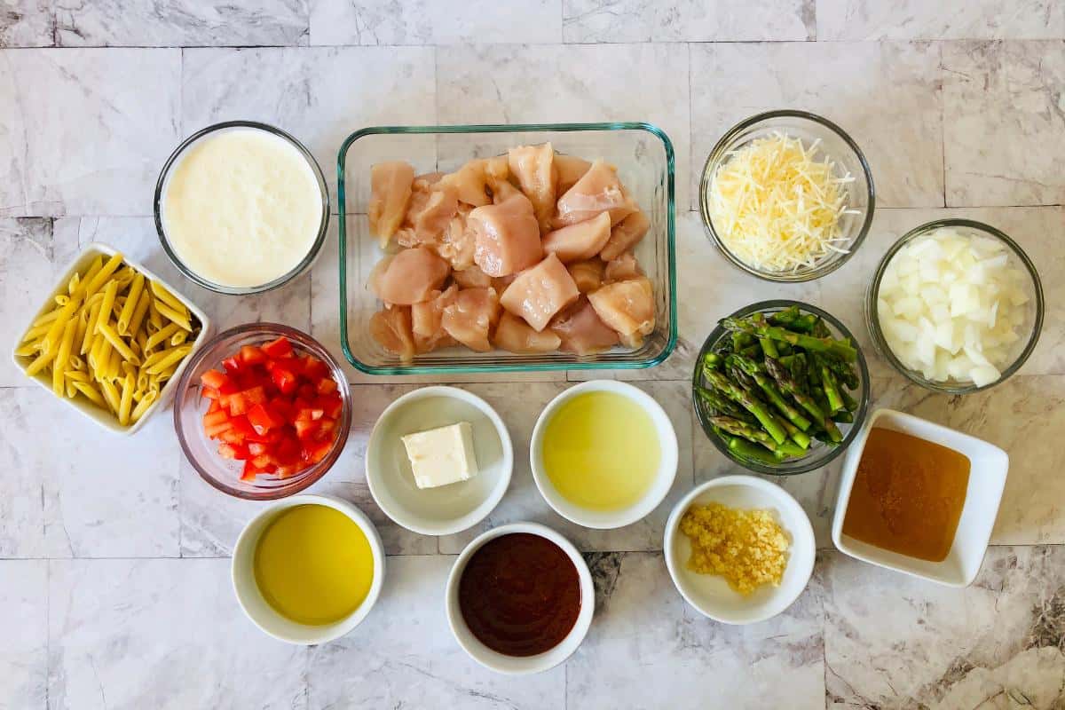 A top-down view of ingredients in bowls for a tasty chicken pasta recipe, including cubed chicken, penne pasta, chopped red bell pepper, asparagus, onion, shredded cheese, cream, butter, grated garlic, oil, and spices.