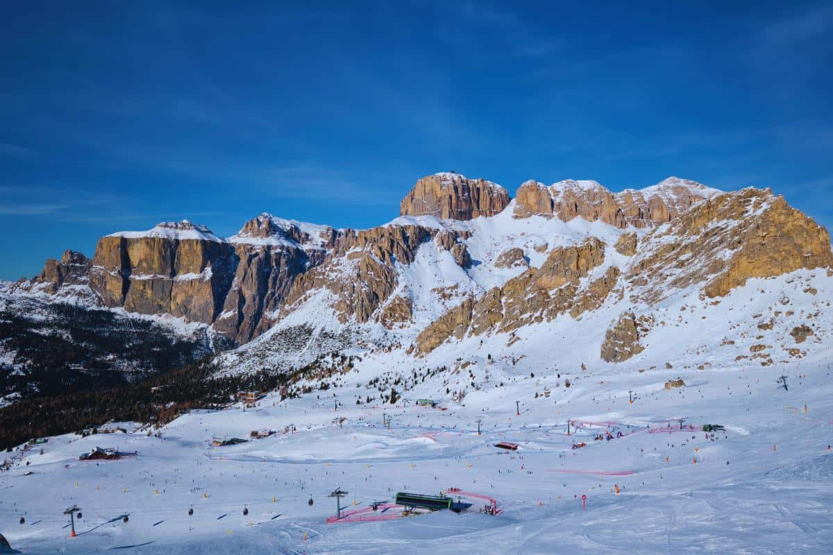 Snow-covered mountains with rugged peaks under a clear blue sky. At the base, ski lifts, slopes, and small buildings hint at a busy Italy ski resort surrounded by rocky cliffs—perfect for those seeking the thrill of skiing in Italy.