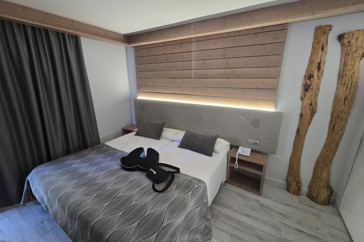 A modern hotel room at Bahia del Sol in Santa Ponsa features a double bed, dark throw pillows, a gray patterned bedspread, and decorative wooden logs. A guitar case rests on the bed, while dark curtains cover the window in this all-inclusive retreat.
