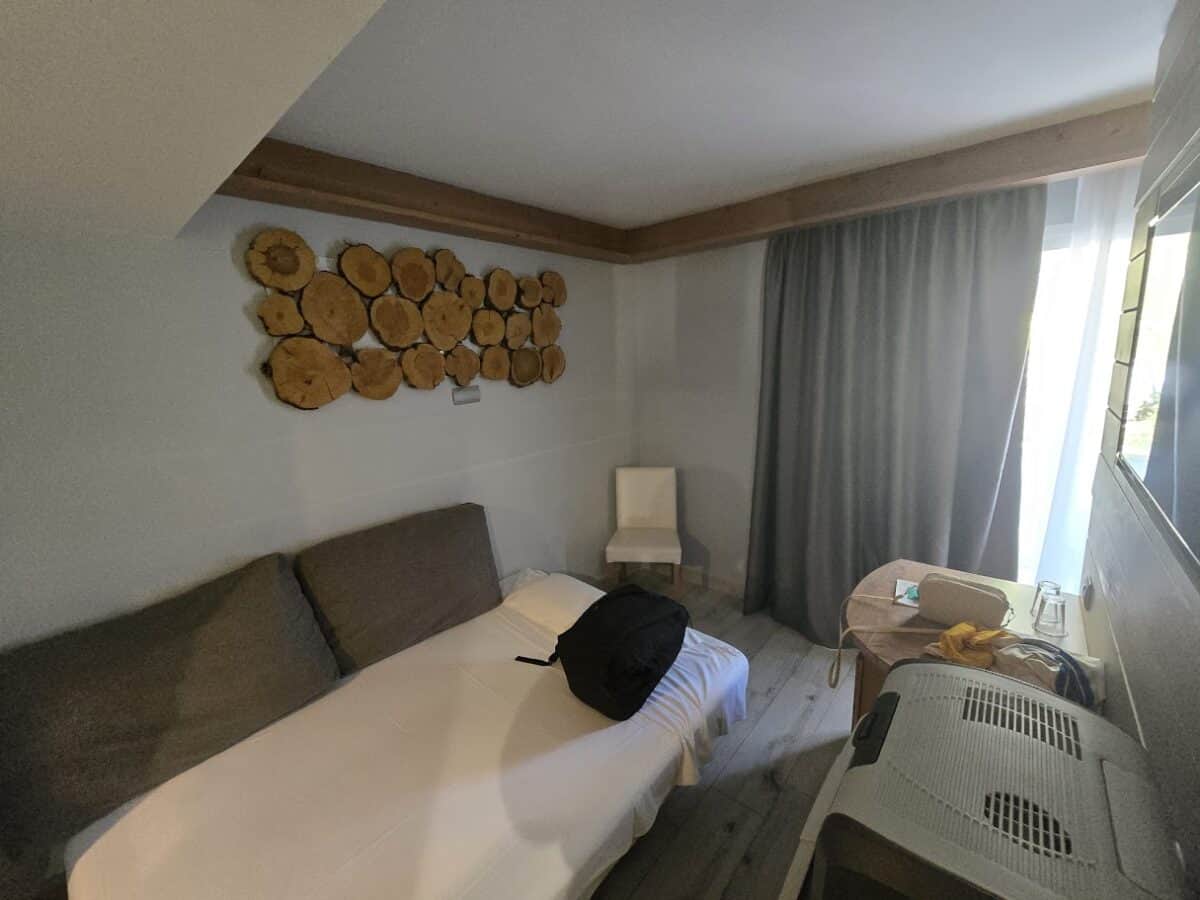 A small hotel room at Bahia del Sol in Santa Ponsa features a single bed with gray cushions, a backpack on the bed, a white chair, round wooden wall art, gray curtains, snacks on a small table, and an air conditioning unit under the wall-mounted TV.