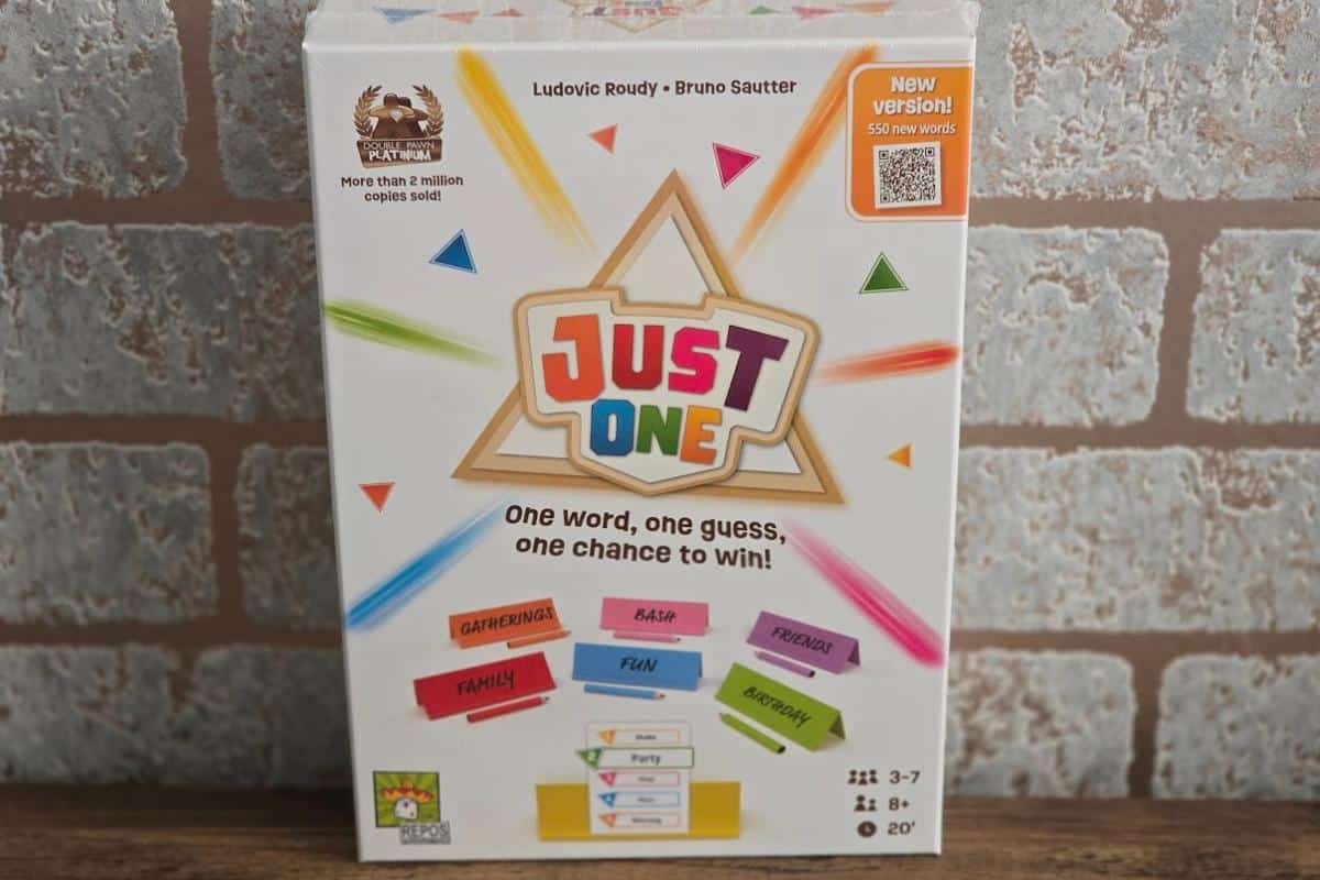 A box of the board game "Just One" stands against a brick wall. The colorful box showcases this lightweight family game’s title and highlights features like “One word, one guess, one chance to win!” and “New version! 550 new words.”.