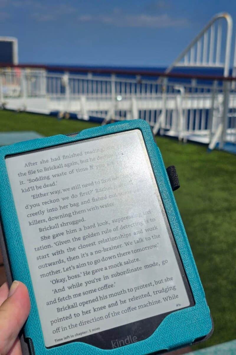 A person holds an Amazon Kindle with a blue cover, displaying a book page, while standing on a grassy deck near white railings. In the background, blue sky and the outline of a Balearia Ferry from Alcudia to Barcelona are visible.