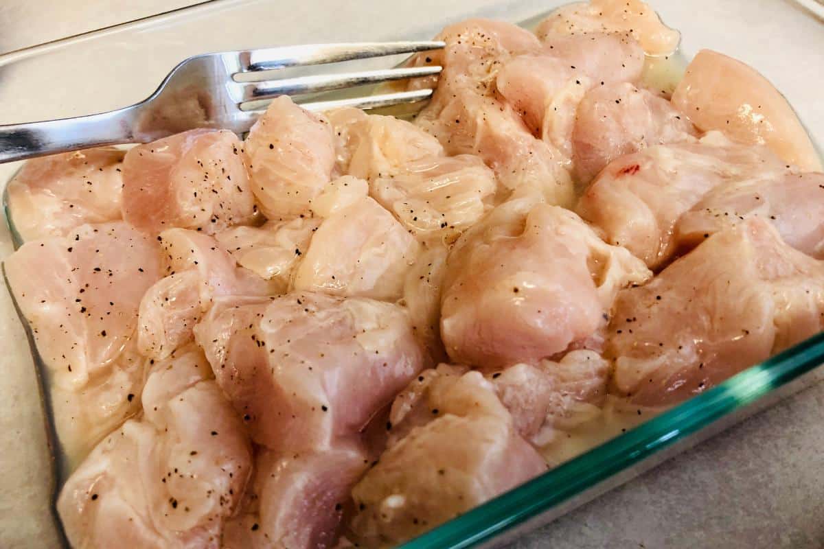 Raw, cubed chicken pieces seasoned with black pepper are arranged in a glass dish, ready to be transformed into a flavorful Chipotle Chicken Pasta, with a metal fork resting on top, ready for cooking.