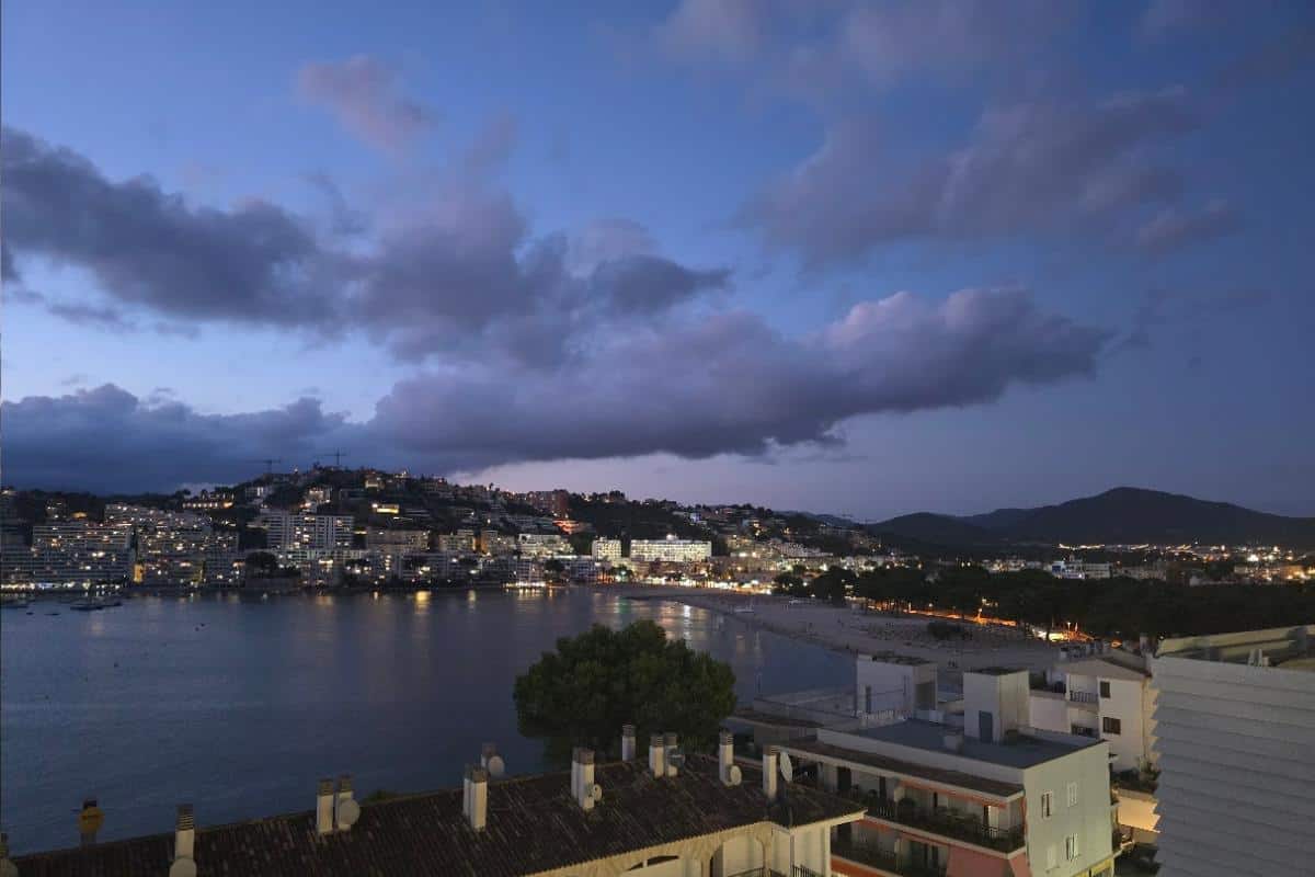 At dusk in Santa Ponsa, lights illuminate buildings along the shoreline of Bahia del Sol. Calm bay waters reflect the glow, while hills rise behind the all-inclusive coastal town beneath scattered twilight clouds.