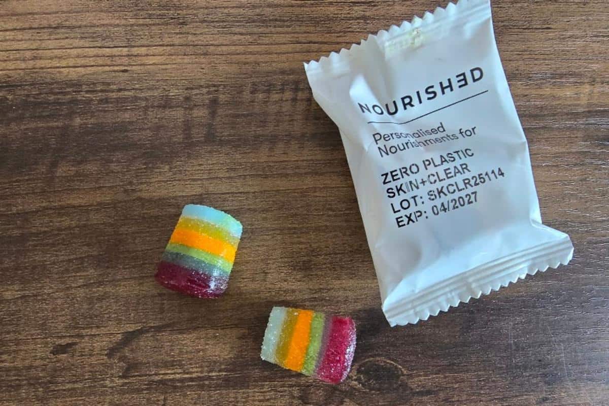 Two colorful, striped collagen gummies and a white packet labeled "NOURISHED Collagen Skin+Clear" lie on a wooden surface. The packet shows an expiration date of 04/2027. An honest review highlights their vibrant look and eco-friendly packaging.