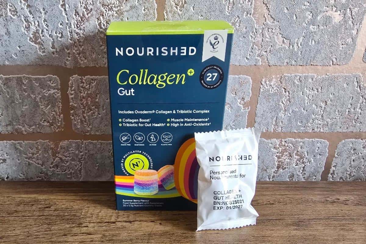A box of Nourished Collagen+ Gut supplements stands on a wooden surface with a single collagen gummy beside it. The box features colorful graphics and brick-patterned wallpaper in the background, perfect for any Nourished Collagen review.