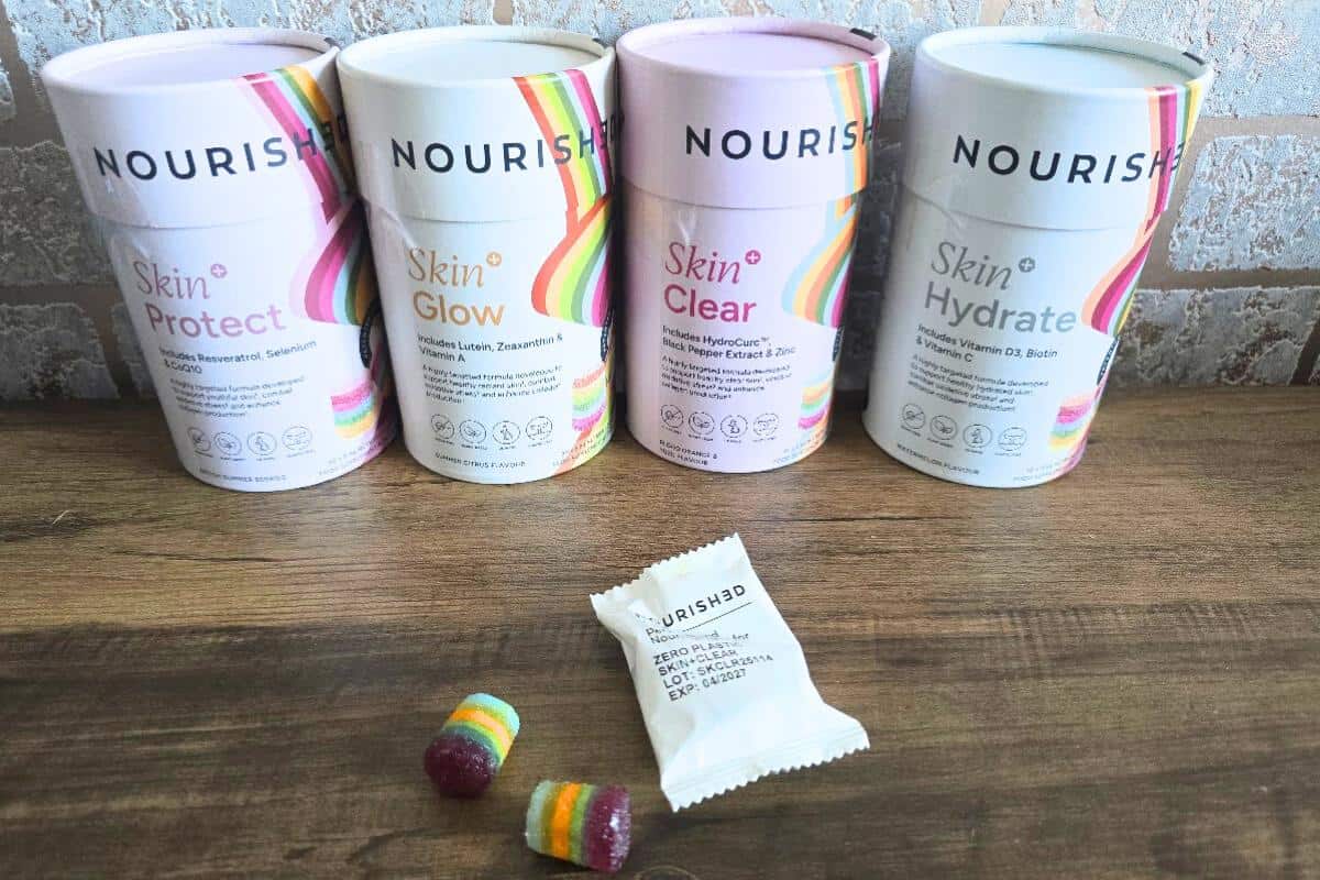 Four cylindrical Nourished Collagen containers (Skin Protect, Skin Glow, Skin Clear, Skin Hydrate) are lined up on a table. In front, there’s a small white packet and two colorful, striped collagen gummies.