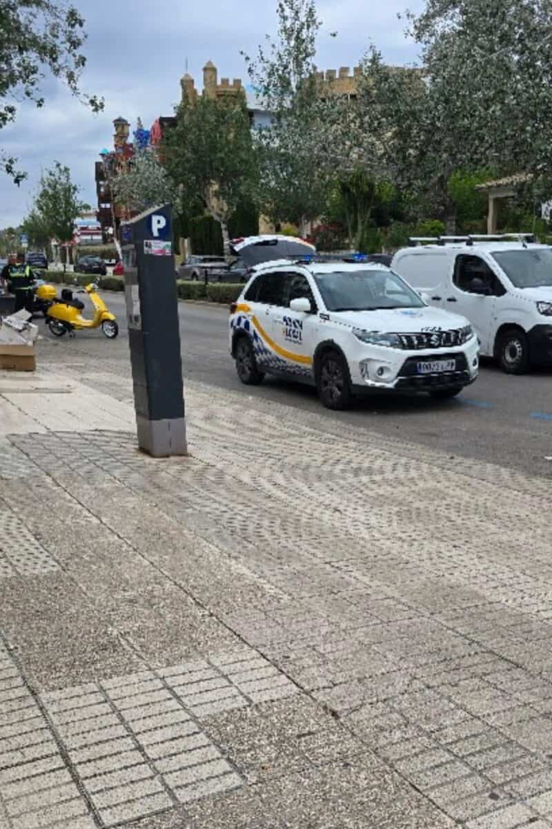 A city street scene perfect for a Mallorca driving guide: a parked traffic police SUV, yellow scooter, and white van. Pedestrians stroll beneath trees and an overcast sky, with a parking meter in the foreground—a glimpse of driving in Mallorca.